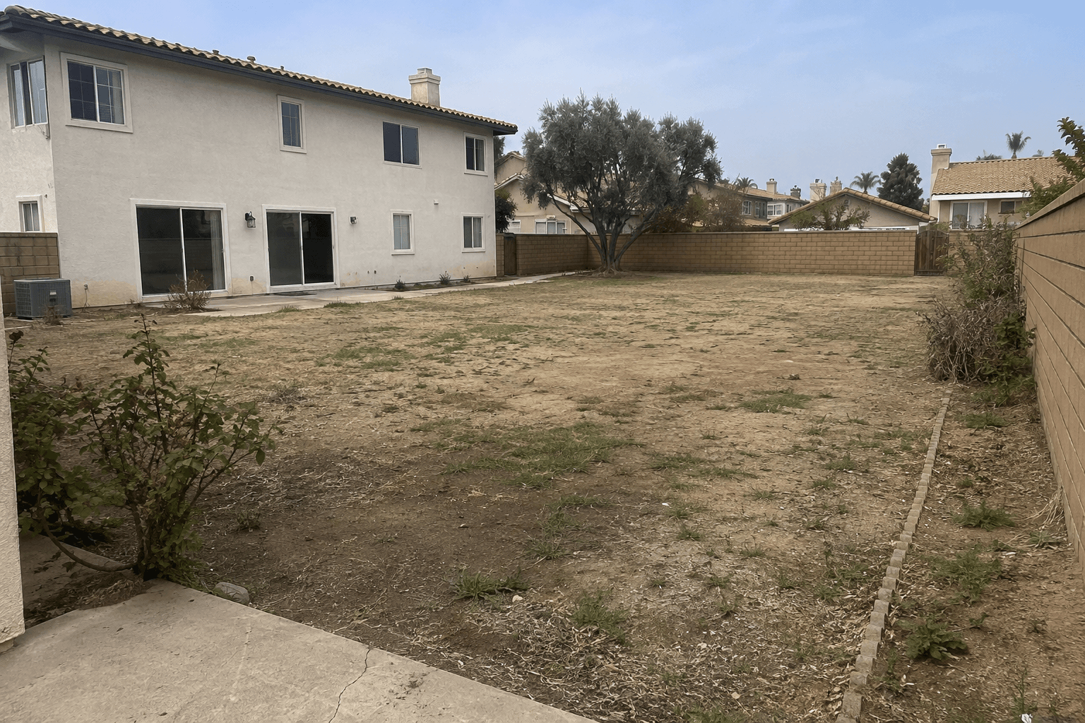 Large unfinished backyard with compacted soil, patchy grass, and a narrow cracked patio before renovation